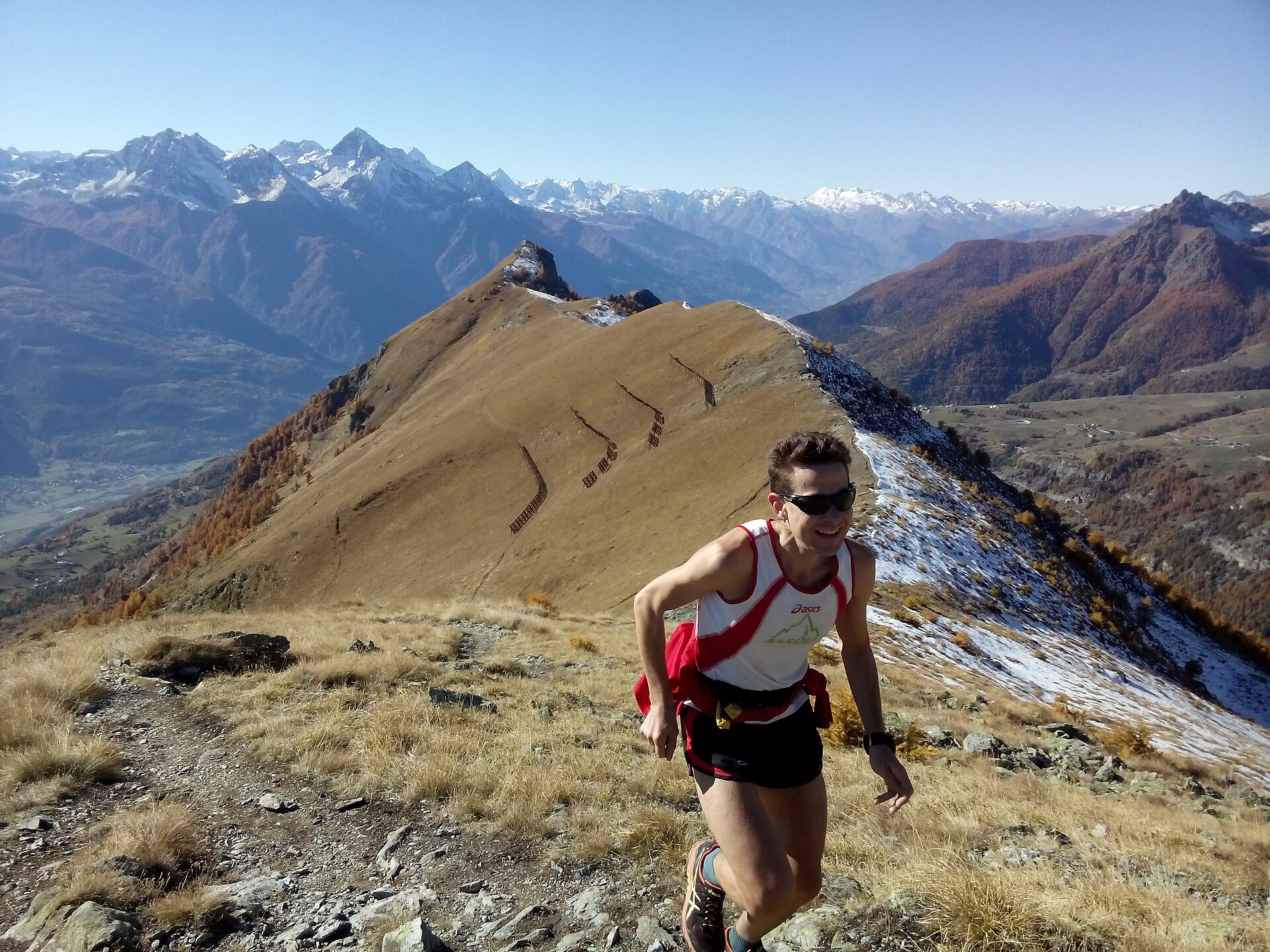 A trail runner climbing a grassy mountain ridge with snowy peaks behind